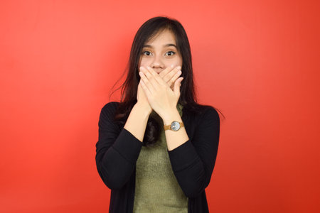 Covering mouth with Shocked Face and Looking at Camera Of Beautiful Asian Woman Isolated On Red...