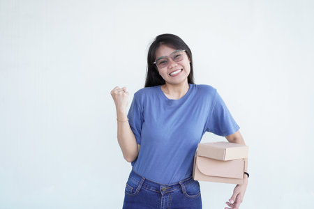 Beautiful Asian woman smiles joyfully, giving a 'yes' gesture while holding a package box against a white backgroundの写真素材
