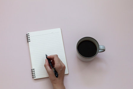 Top view of female hand writing on empty notebook near cup of black coffee on pink background