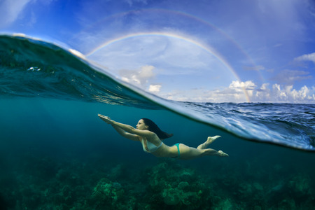 Underwater Sport Postcard. A freediver floating under water surface in ocean. A rainbow appear on cloudy sky over beautiful seascapeの写真素材