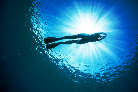An ocean Water surface with bright sun underwater behind ripples. Silhouette of female freediver floating through sunbeams.の写真素材