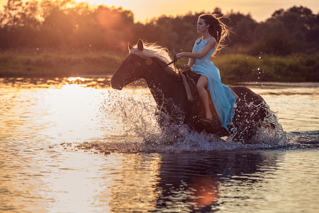 Girl riding horse in a river on the background of sunset sky and glossy water. Female model wearing blue dress on horseback at sunset time, backlit with sunshine with water drops and splashesの写真素材