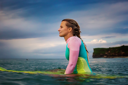 Surfer girl in bright swimsuit waiting for the wave in open water. Sunset time at the ocean. A woman sitting on surfboard in water.の写真素材