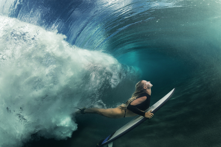 A blonde surfer girl underwater doing duck dive holding surfing board left behind air bubbles in blue water background under big ocean waveの写真素材