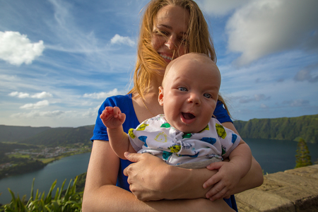 A four months old little baby on mothers hands wondering with joy outdoors. A woman wearing blue dress standing against beautiful landscape on background.の写真素材