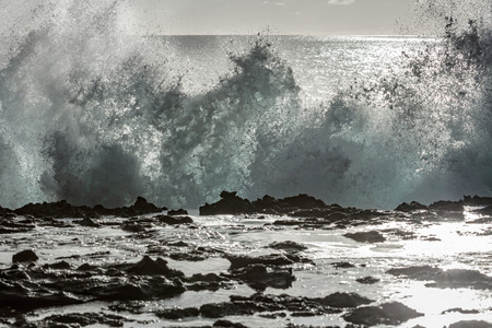 Shorebreak water splash. Ocean wave breaking against black volcanic rocks. Bright sunlight at backgroundの写真素材