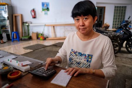 Asian Indonesian women, the owner of small local family-owned business store, or locally called warung, calculating profit by her desk. Selective Focus.の写真素材