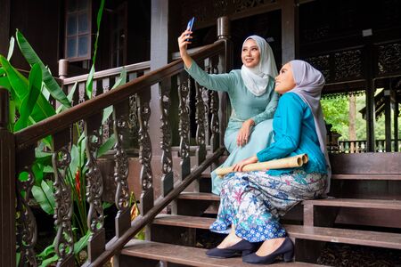 Muslim Malay women wearing hijab and traditional costume are taking selfie during Aidilfitri celebrations at the terrace of traditional wooden house. Hari Raya and Muslim women fashion attire concept.の写真素材