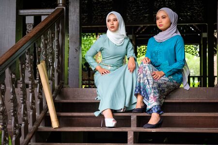Portrait of Muslim Malay women wearing hijab and traditional costume during Aidilfitri celebrations at the terrace of traditional wooden house. Hari Raya and Muslim women fashion attire concept.の写真素材