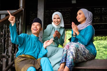 A group of three malay muslim people in traditional costume are having happy conversation during Aidilfitri celebration at terrace of traditional wooden house. Raya and Muslim fashion attire concept.の写真素材
