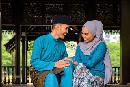 Young couple of malay muslim in traditional costume having happy conversation during Aidilfitri celebration at traditional wooden house. Raya and Muslim fashion attire concept.の写真素材