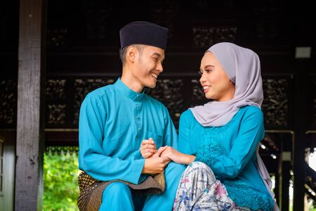 A portrait of young couple of malay muslim looking each other in traditional costume during Aidilfitri celebration at the traditional wooden house. Raya and Muslim fashion attire concept.の写真素材