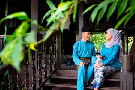 A portrait of young couple of malay muslim having conversation in traditional costume during Aidilfitri celebration at the traditional wooden house. Raya and Muslim fashion attire concept.の写真素材