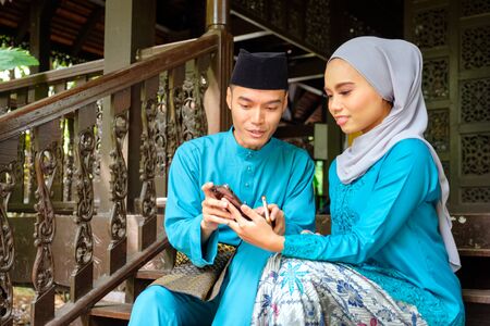 Young couple of malay muslim in traditional costume watching online content in a smart phone during Eid al-Fitr celebration at wooden stair. Raya and Muslim fashion attire concept.の写真素材