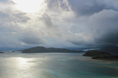 Dramatic Tropical Island Landscape Under Stormy Sky with Sunlight Reflections on Ocean Surfaceの写真素材