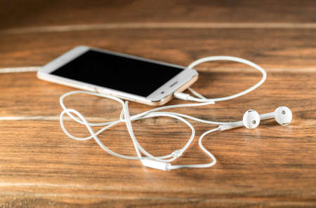 A smartphone with white wired headphones on a wooden table