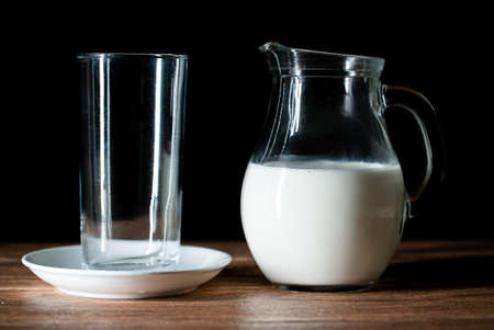 An empty glass cup and a jar of cow's milk on a wooden table with black backgroundの写真素材