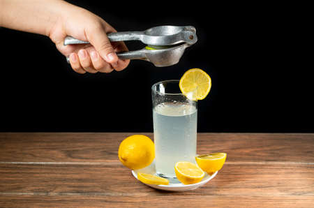 Squeezing a lemon into a lemonade glass with yellow lemons around a white plate, making a lemonade dark foodの写真素材