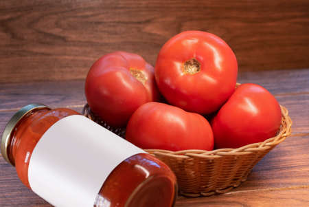 many red tomatoes in a woven basket with a glass of tomato paste on a wooden boardの写真素材