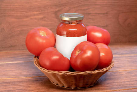 many red tomatoes in a woven basket with a glass of tomato paste on a wooden boardの写真素材