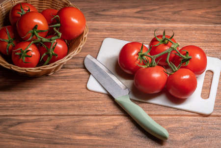Chopping board with ripe tomatoes and stainless steel knives on left side plus basket full of fresh...