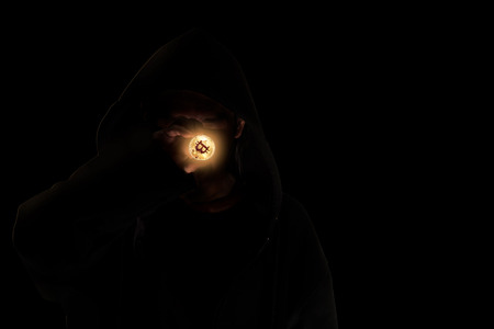 Hooded man in shadow holding crypto currency bitcoin on blabk background.の写真素材