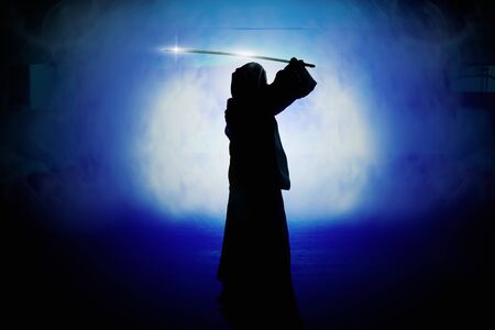 Silhouette of man in robe holding sword with smoke and light in parking lot.