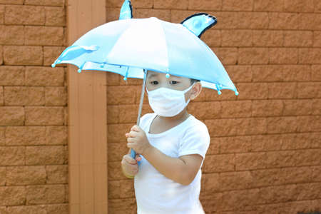 Little Asian boy holding an umbrella and wearing face mask protection from influenza and coronavirus illnesses.の写真素材