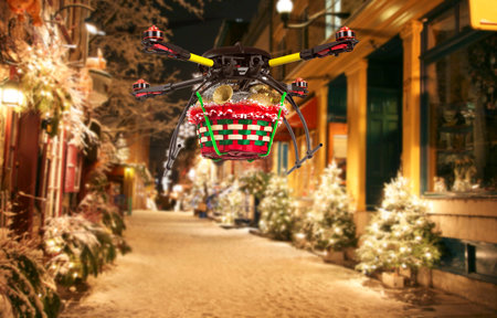 A flying quadrocopter delivering a basket of Christmas goodies above covered with snow beautifully decorated streetの写真素材