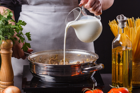 young woman in a gray apron adds milk to mushroom sauce.の写真素材