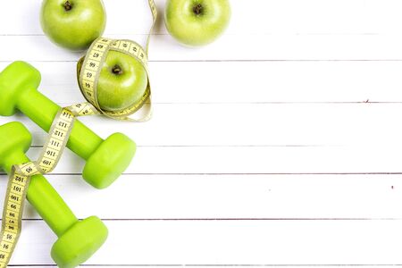 Green dumbbells and green apples with measuring tape and copy space on white wooden background. Sport, fitness and diet concept. Top view.の写真素材