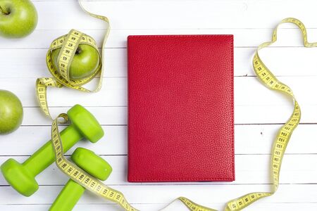 Red notepad with green dumbbells and apples with measuring tape on white wooden background. Sport, fitness and diet concept. Top view.の写真素材