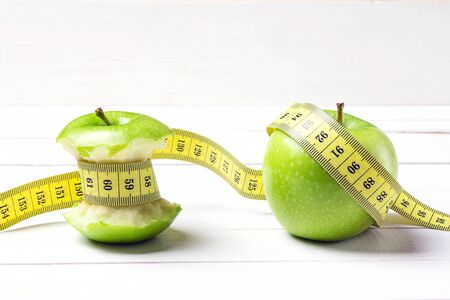Green apple and apple stump with yellow centimeter tape on white wooden background. Concept sport, diet, fitness, anorexia.の写真素材