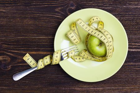 Diet concept represented by fresh green apple on plate with yellow measuring tape knotted around and  fork on wooden table.  Weight loss and healthy.の写真素材