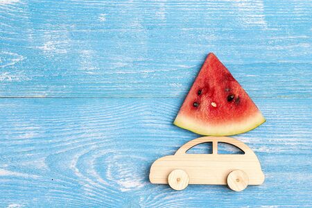 Wooden toy car with slice of watermelon on the roof on blue background. Copy space for text, top view.の写真素材