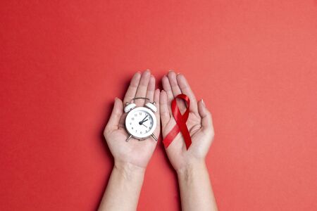 Female hands holding red AIDS awareness ribbon and alarm clock on red background. 1 december World...