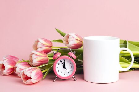 White coffee mug with tulip flowers and  alarm clock on pink  background. Space for text or design....