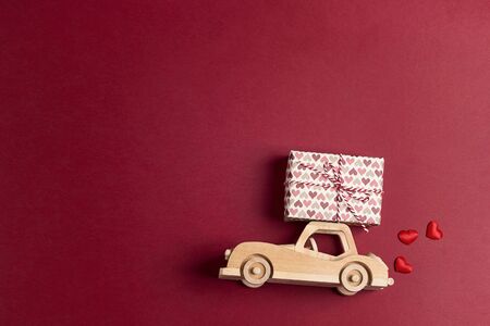 Wooden toy car with a gift box on the roof on red background. Copy space for text. Valentine's day...
