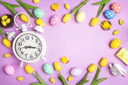 Easter alarm clock with colorful eggs and tulips on purple background. Copy space. Time to celebrate.の写真素材