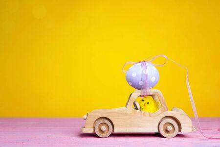 Wooden toy car with Easter egg on the roof and chick driver on yellow background. Copy space.の写真素材