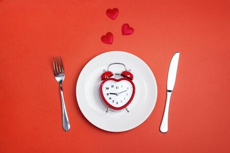 Festive table setting with cutlery and heart-shaped alarm clock on red background. Top view. Time...