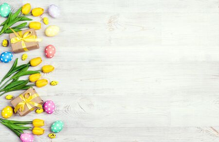 Easter border from colored eggs, gifts and tulips on light wooden background.  Top view, space for...