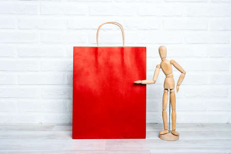 Red paper shopping bag with wooden dummy in a proposing pose on white brick wall background. Copy space for text. Shopping, sale, Black Friday concept.の写真素材
