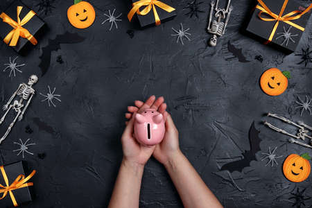 Female hands holding a piggy bank surrounded by halloween decorations on black background. Save money to celebrate Halloween.の写真素材