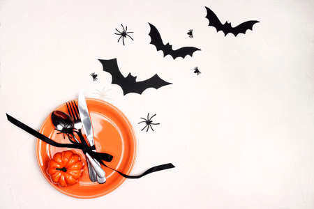 Halloween table setting with orange plate, cutlery and spooky decorations on white table. Top down...