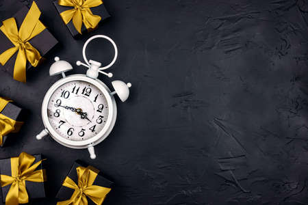 White alarm clock surrounded by black gift boxes with gold ribbons on a black background. Holiday or...