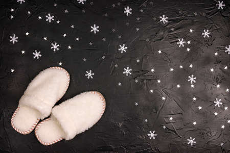 White fluffy house slippers on a black background with stars and snowflakes. Home winter coziness concept.の写真素材