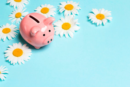 Pink piggy bank with chamomile flowers on a blue background. Copy space for text. Save up money for the summer.の写真素材