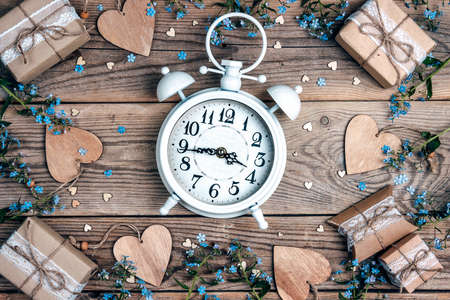 White vintage alarm clock with forget-me-not flowers, gifts and hearts on old wooden boards.