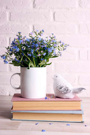 White coffee mug with bouquet of forget-me-not flowers, books and bird on the white brick wall background. Space for text or design.の写真素材